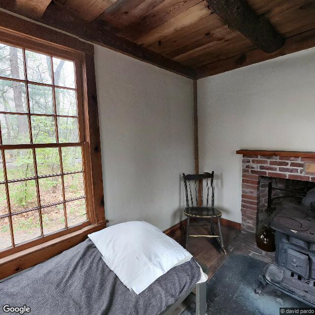 Inside the replica of Thoreau’s cabin: cot, wood stove, chair, one window. Street view © Google, photo credit david pardo.